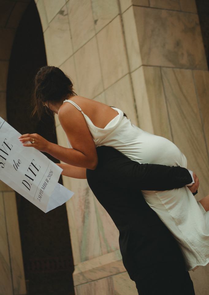 He lifts her against a stone archway, save-the-date in hand